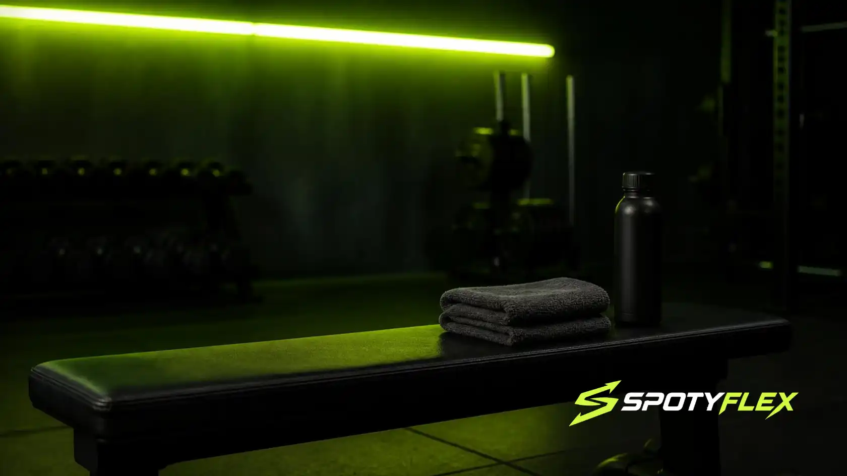 An empty flat bench with a towel and water bottle in a dark gym, lit by a neon green light bar